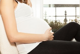 Eine schwangere Frau mit weißem Hemd und schwarzer Hose sitzt auf einem Sofa und hält ihren Bauch von unten fest Kopf und die kompletten Beine sind zu sehen / Schwangere Frau